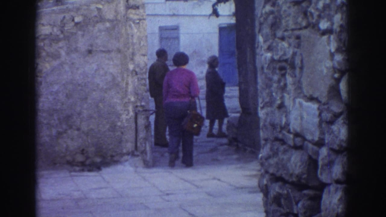 Several adults stand or walk near a bend in a stone alley with a blue door beyond.