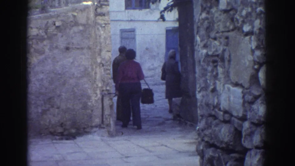 1976 Tel Aviv frame showing a woman with a handbag and other adults paused in a weathered stone alley.