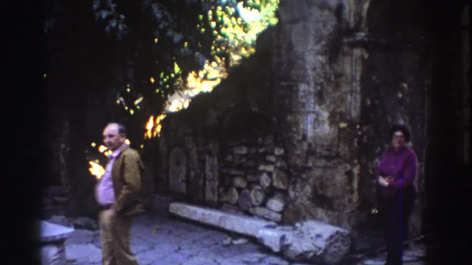 1976 Tel Aviv home-movie frame of a man and woman standing in a shaded stone alley area with trees overhead.