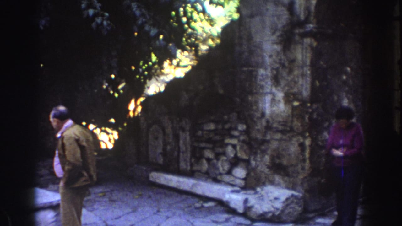1976 Tel Aviv home-movie frame showing an older man and woman near stone walls, arched recesses, and tree shade at ruins.