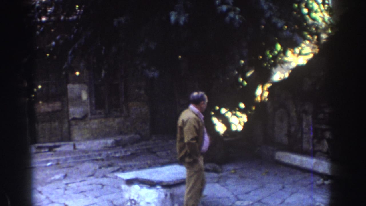 Older man walking across a stone courtyard beside weathered walls and trees.