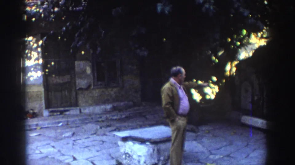 1976 Tel Aviv home-movie frame of an older man standing among stone paving, a low slab, and old masonry in shade.