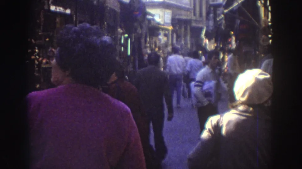 A handheld view follows pedestrians through a narrow Tel Aviv market passage, with shop displays and bright daylight ahead.