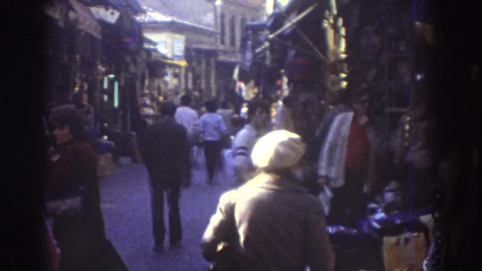 The final frame looks down the busy 1976 Tel Aviv market lane, with pedestrians, hanging merchandise, and strong daylight at the far end.