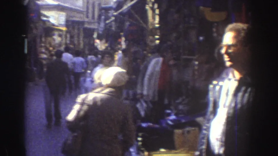 1976 Tel Aviv market street with pedestrians moving through a narrow shopping lane lined with storefronts and hanging goods.