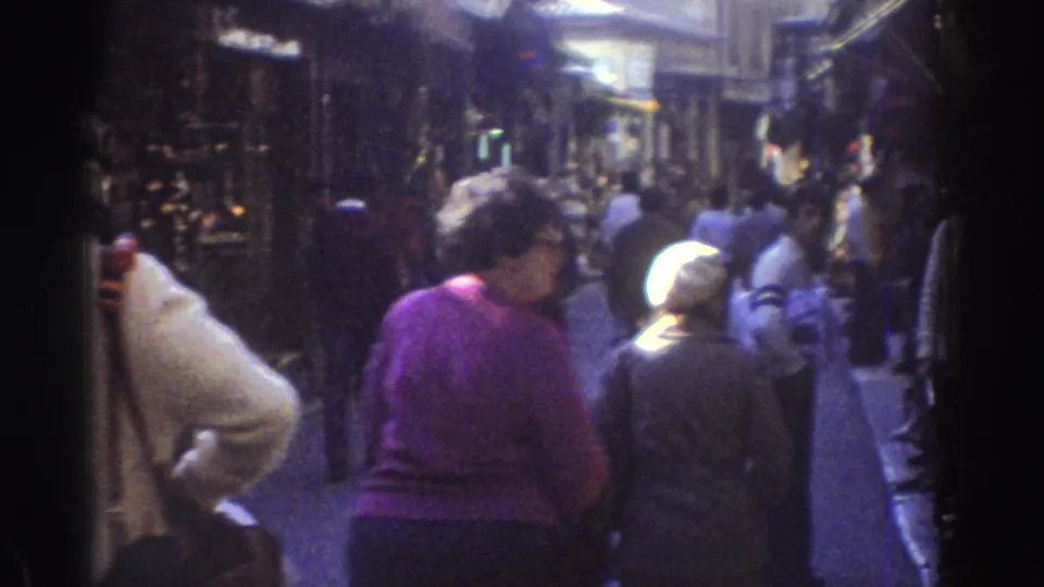 People in 1970s street clothing move through a Tel Aviv shopping lane, with stalls and storefronts filling the background.