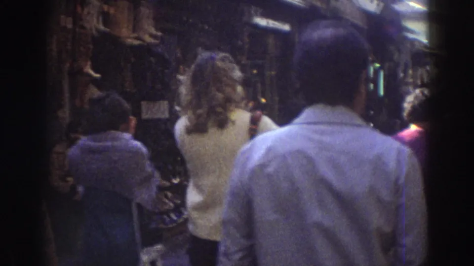Handheld 8mm view from behind several pedestrians walking through a shaded Tel Aviv market street in 1976.