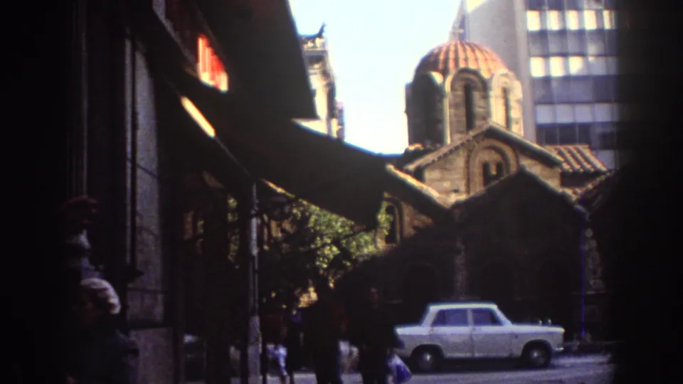 A handheld 1976 Tel Aviv frame shows a red-domed historic church-like building, pedestrians, and a white sedan from a shaded sidewalk view.