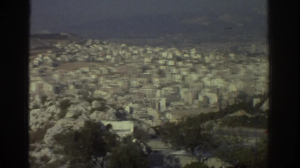 1976 Tel Aviv cityscape with soft-focus buildings, rocky foreground, scattered trees and muted 8mm color.
