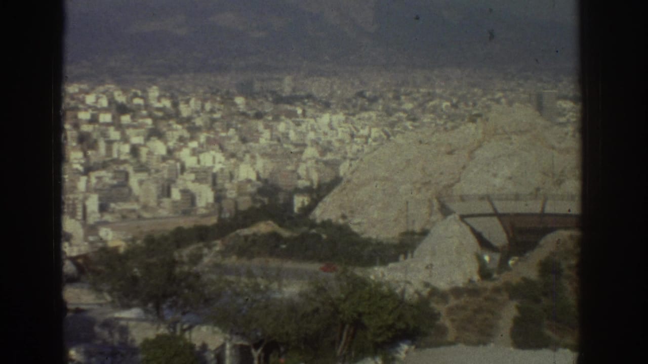 1976 Tel Aviv home-movie frame showing a high view over dense buildings, pale rocky hillside, trees, and distant hills.