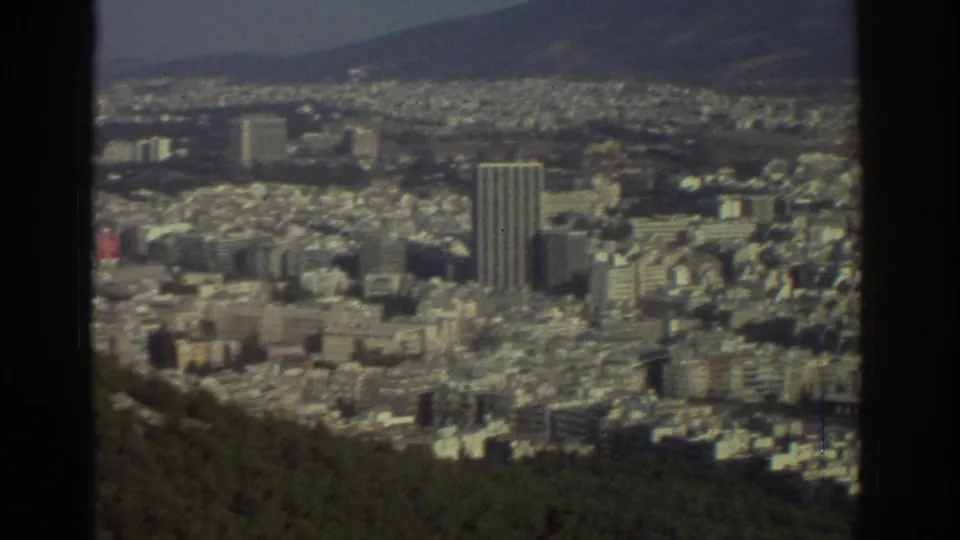 1976 Tel Aviv home-movie view with a prominent high-rise tower among lower buildings, wooded hillside foreground, and distant hills.