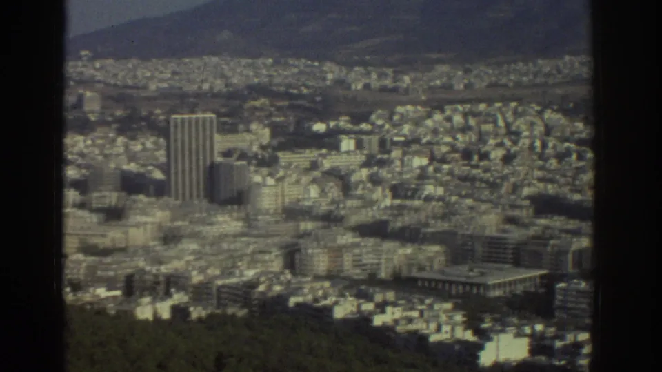 1976 Tel Aviv frame showing the continued pan across dense city blocks, a tall tower, hillside greenery, and distant terrain.