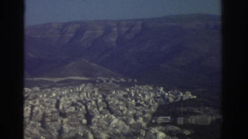 Soft, grainy 1976 Tel Aviv elevated view showing a broad city area below hills and dark frame edges from the shooting position.