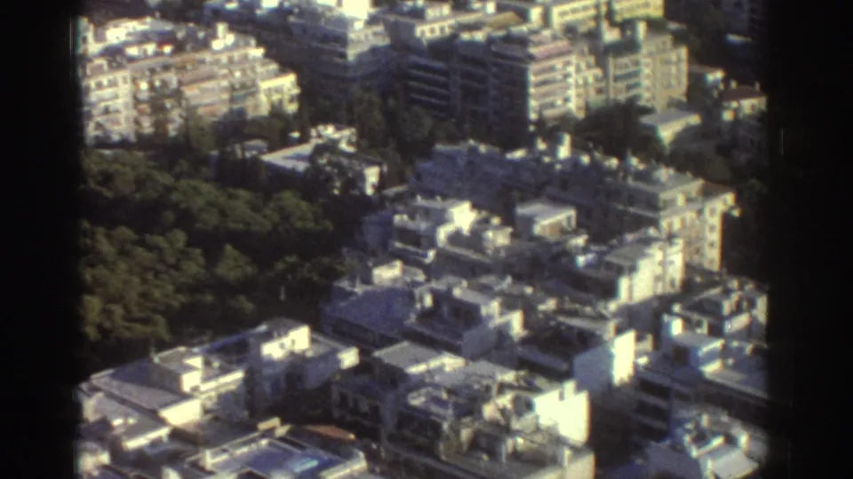 Tel Aviv residential cityscape from above, with 1970s 8mm softness, bright rooftops, apartment blocks, and tree cover.