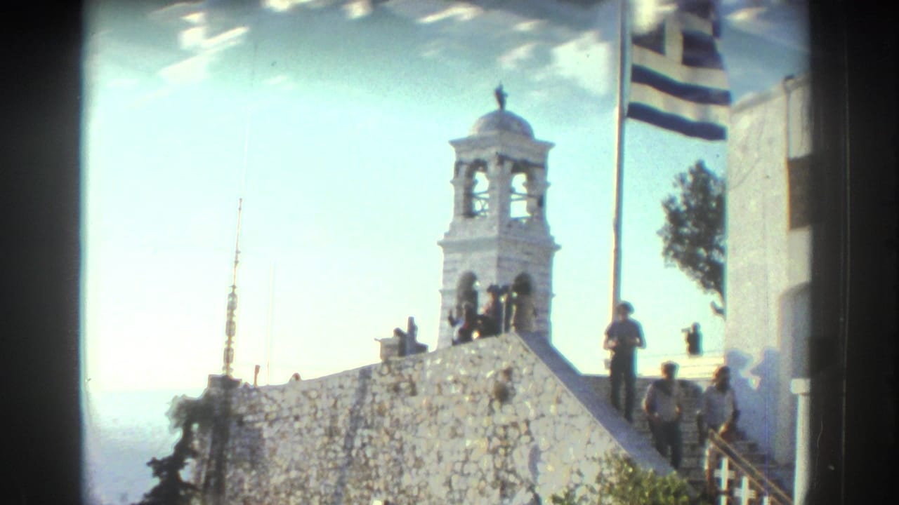 White bell tower above a stone wall with a Greek flag and people on steps.
