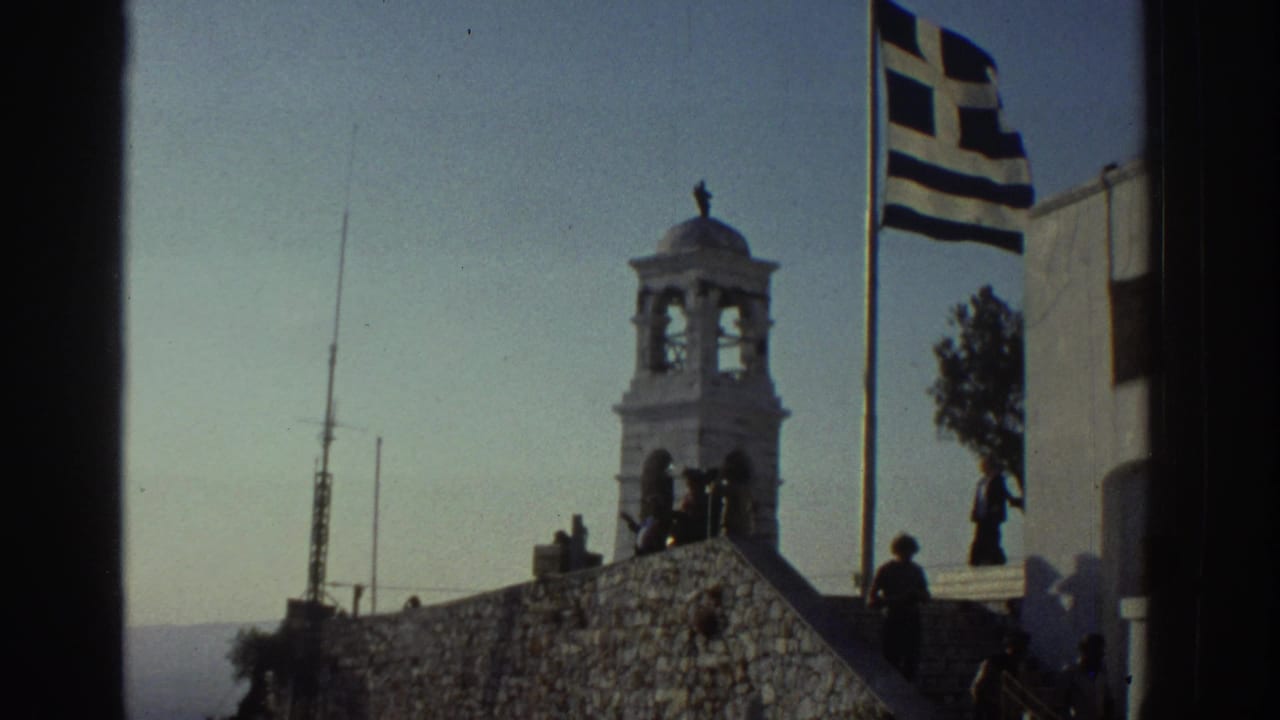 Bell tower and Greek flag beside a sunlit stone terrace with silhouetted people.