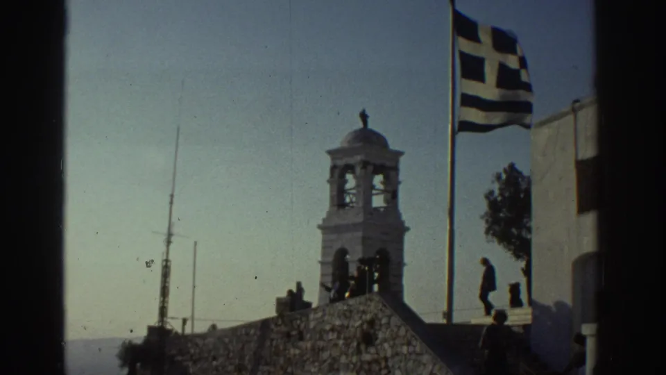 1970s 8mm frame showing the Greek flag near a white building, bell tower, stone wall, and a lone pedestrian.