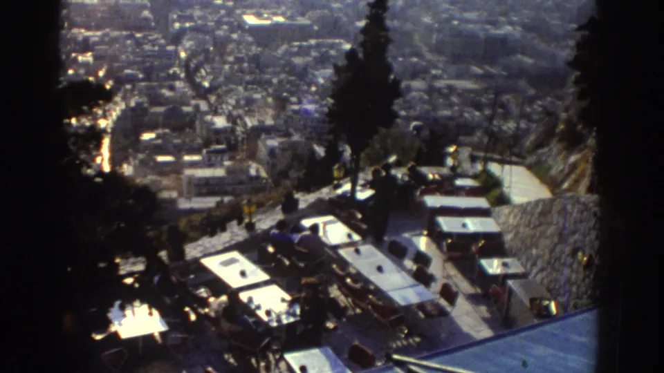 1970s 8mm overlook from a hillside terrace, with tables, trees, stone walls, pedestrians, and a distant urban panorama.