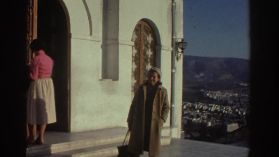 1976 Israel home-movie frame showing two women on church steps beside a white facade and distant city view.