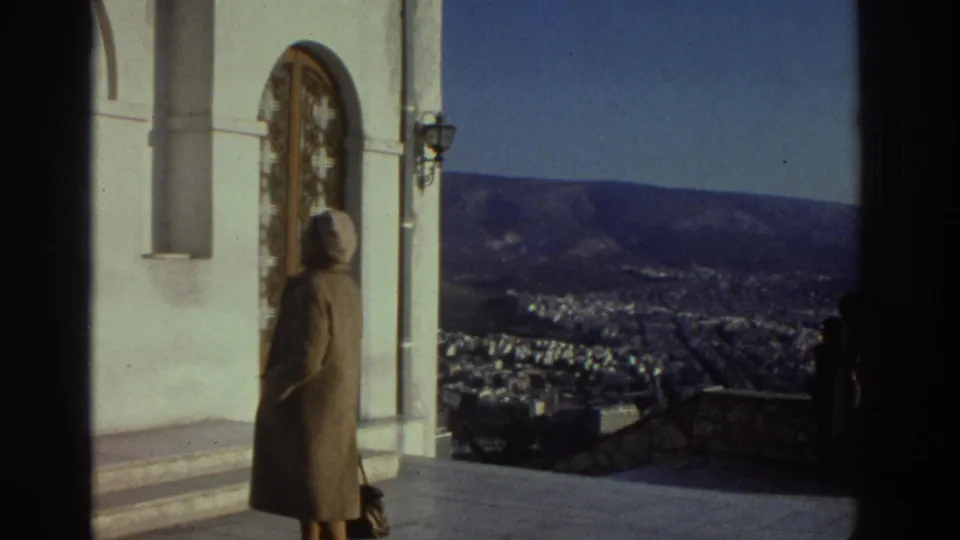 1976 Israel home-movie frame showing a woman in a coat and headscarf near an arched church doorway with a city and hills below.