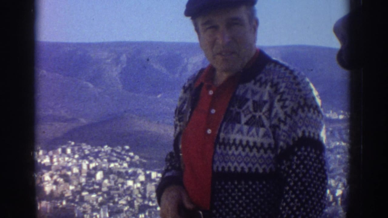 1976 Israel travel footage frame with a smiling man in a patterned cardigan and cap standing against a sunlit city view.