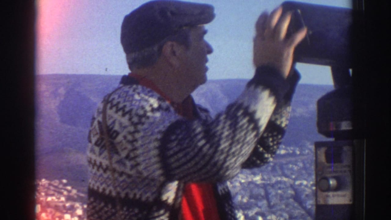 Man in a cap reaches toward a coin-operated binocular viewer at an overlook.