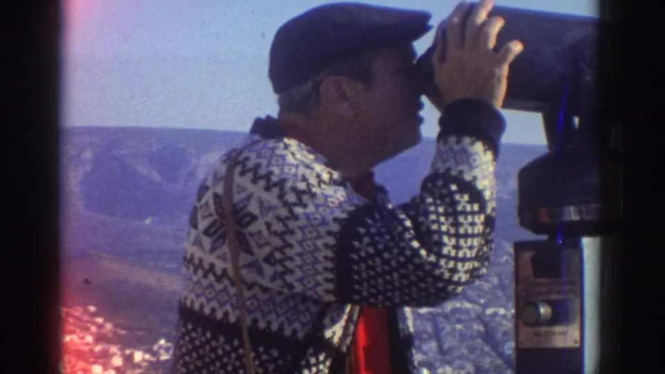 A man in a patterned sweater peers into a coin-operated viewer, with the Tel Aviv-area cityscape and hills visible in the background.