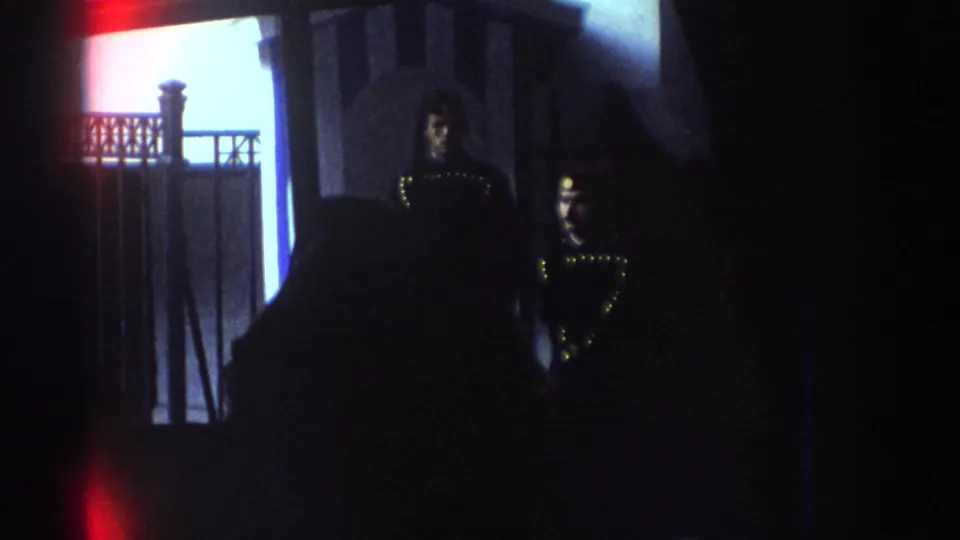 A dim interior shot shows two ornate uniformed figures near a doorway or display area, filmed with strong shadow and home-movie exposure shifts.