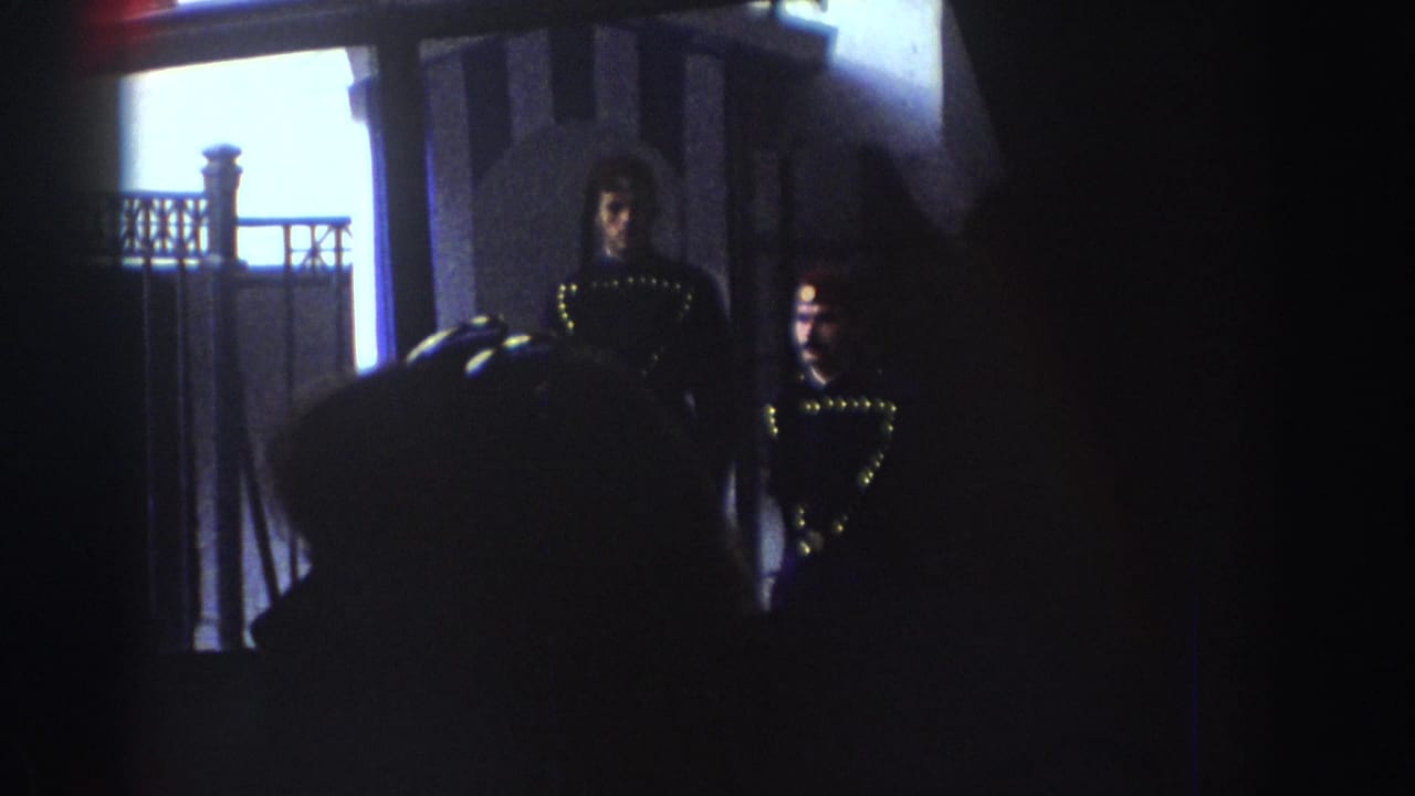 1970s 8mm frame with two visible people in shadow near a doorway or balcony, partly obscured by dark film edges.