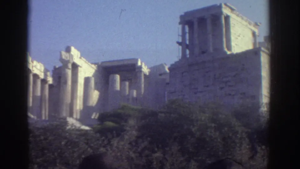 1976 8mm view of pale stone columns and ruin walls, with foreground vegetation and a dark vertical frame edge.