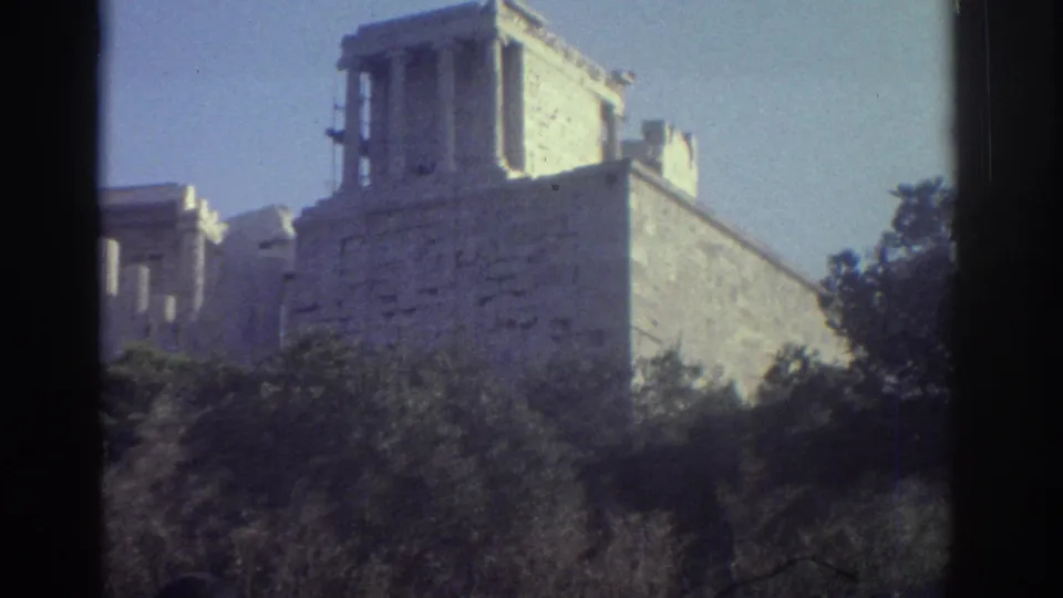 Metadata-supported 1976 home-movie frame of classical stone ruins rising above shrubs, with soft exposure and visible film borders.