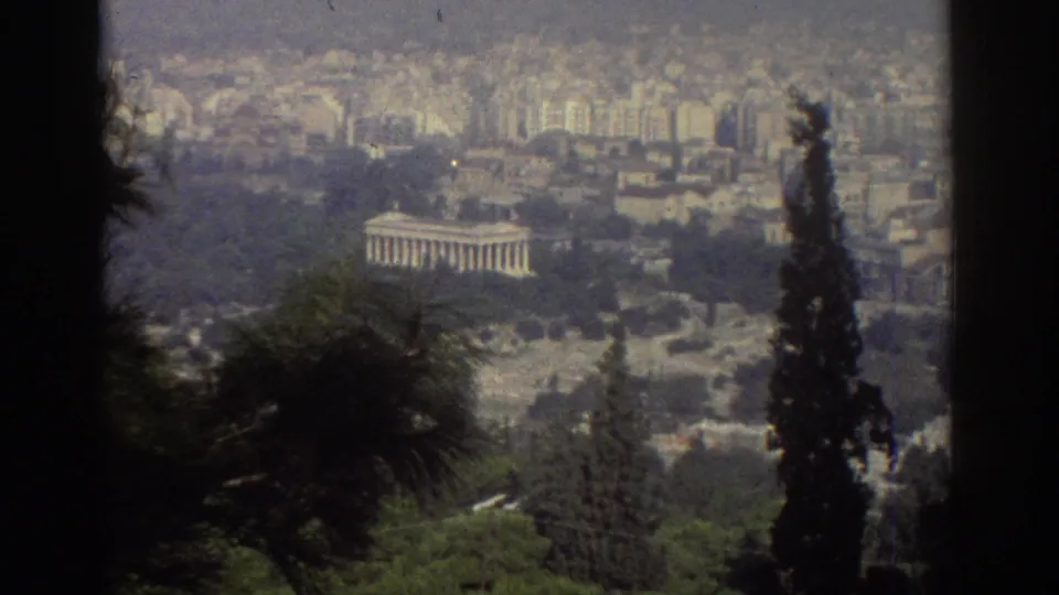 1970s 8mm travel frame showing a distant classical-style columned building, trees, and a hazy urban backdrop.