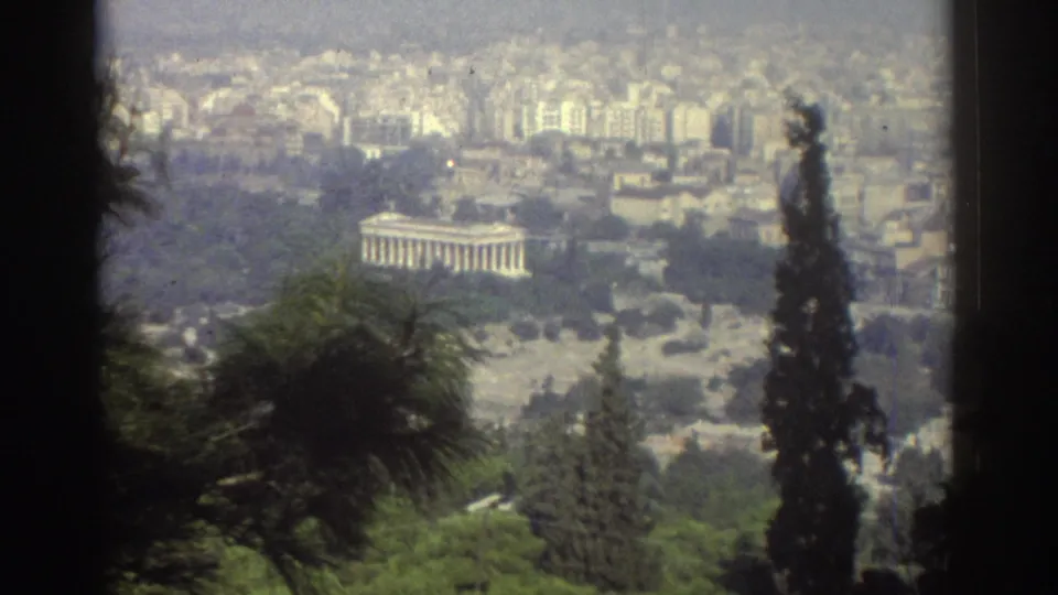 1970s 8mm home-movie view across trees toward a columned monument and surrounding urban buildings.