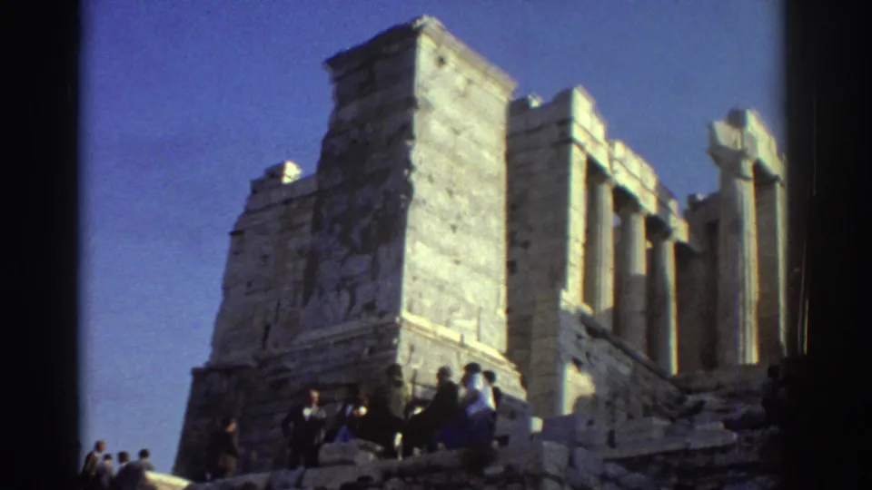 1970s 8mm travel frame of tourists among large pale stone ruins, with columns and steps visible under clear sky.