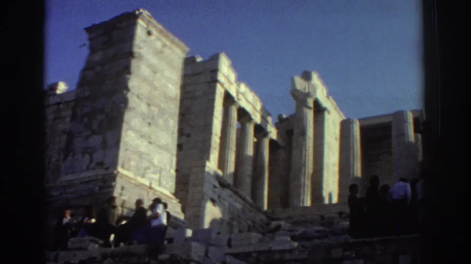 1970s 8mm home-movie frame showing visitors at monumental classical ruins with tall columns, stone walls, and deep shadows.