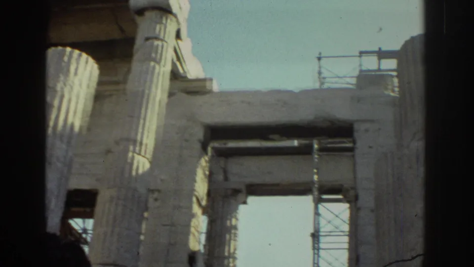 Handheld 8mm frame with visitor silhouettes below fluted columns, a stone opening, and restoration scaffolding against the sky.