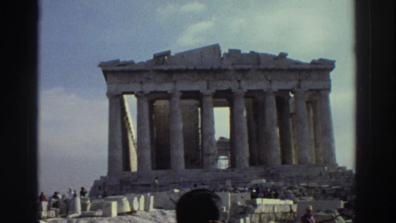 A columned ancient ruin appears under cloudy sky as tourists gather around rubble and stone blocks in the foreground.