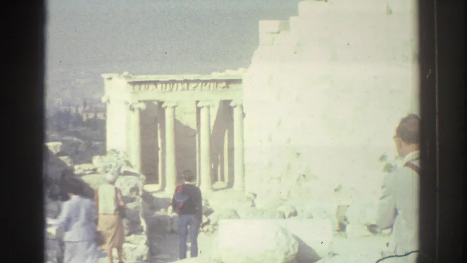 Visitors in casual clothing stand among pale stone rubble near columned ruins, with a hazy city view beyond the site.