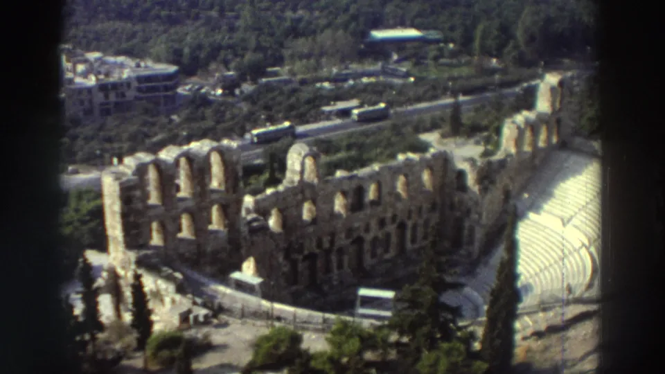 1976 travel footage frame showing a stepped amphitheater beside greenery, roads, and parked buses.