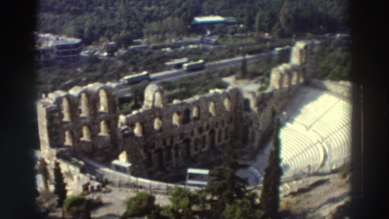 1976 8mm travel frame showing an elevated view of the Odeon of Herodes Atticus ruins, with arched stone walls, curved seating, trees, roads, and buses.