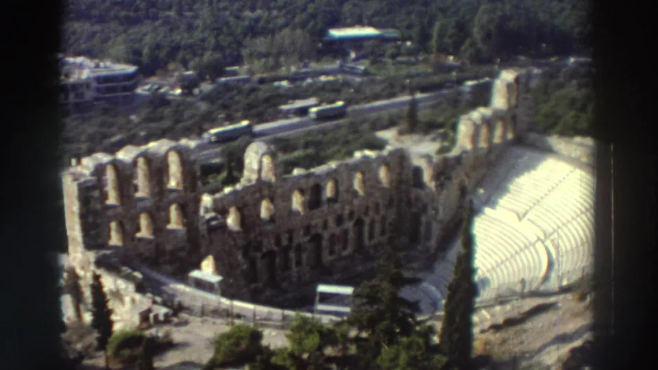 1976 8mm travel frame showing an elevated view of the Odeon of Herodes Atticus ruins, with arched stone walls, curved seating, trees, roads, and buses.
