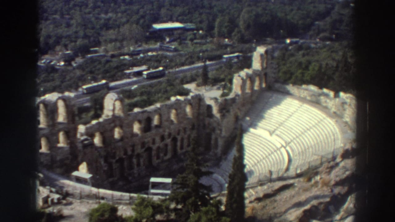 Similar elevated view of amphitheater ruins with pale curved seating, arched masonry, roads, buses, and wooded hillside.