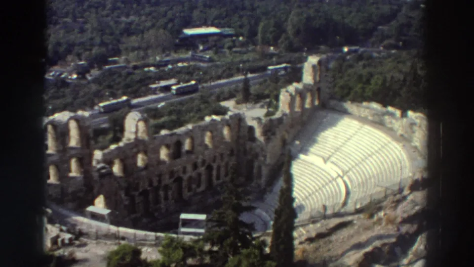 1976 8mm frame with an elevated tourist view of the ancient amphitheater, showing curved seating, arched stone walls, roads, buses, and trees.