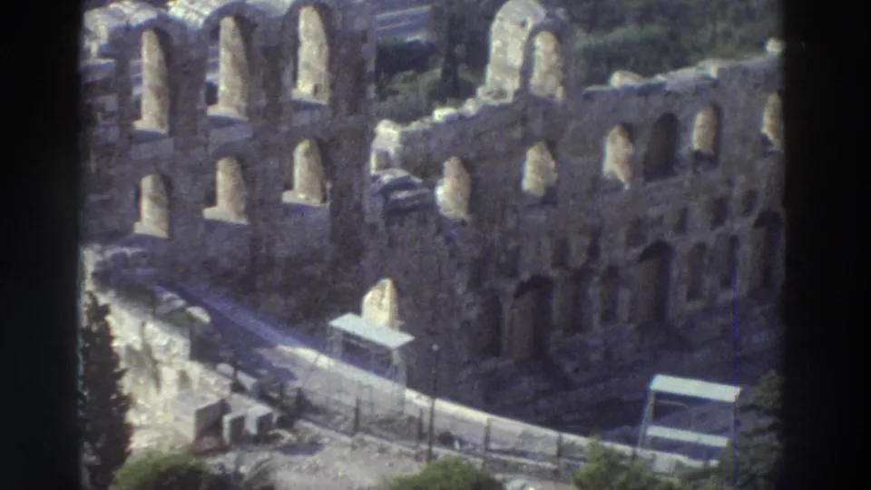 Closer 1976 8mm view of the amphitheater's arched stone wall, with pathways, railings, small sign structures, trees, and soft home-movie focus.