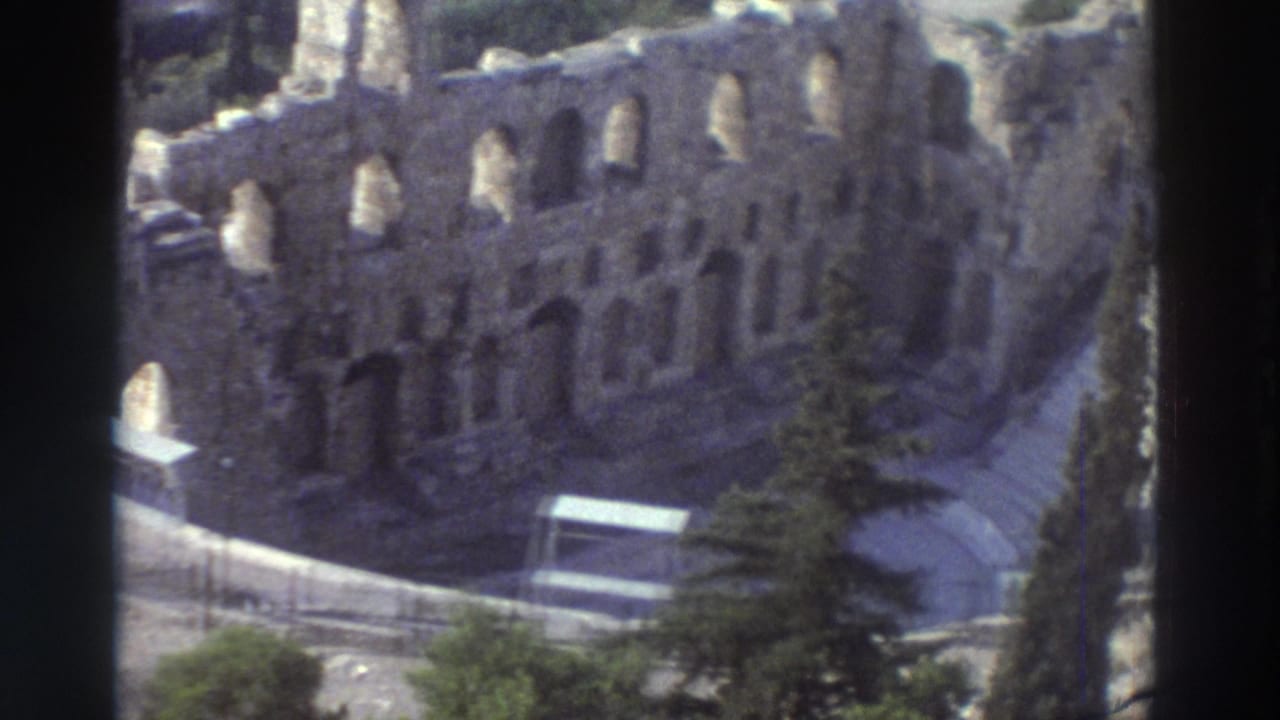 Elevated view of arched stone ruins beside curved stadium seating and cypress trees.