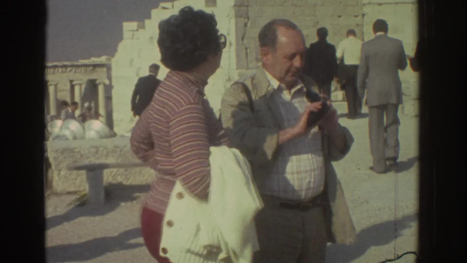 1976 tourist home-movie frame showing a man with a still camera and a woman near pale ruins, with other visitors in the background.
