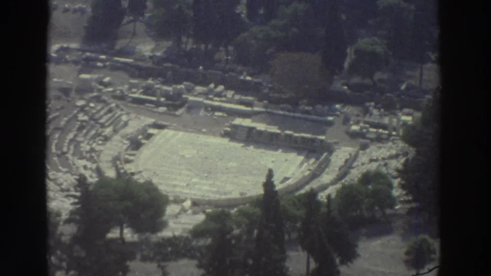 Final 8mm frame holds on the archaeological site from above, showing pale stone seating, ruins, and trees around the arena.