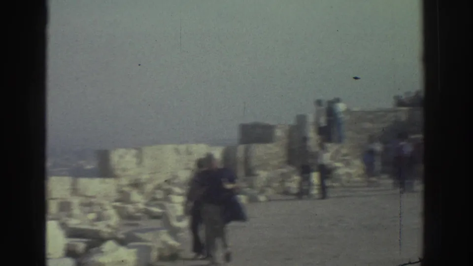 Visitors in a 1976 travel-film scene walk through a hazy archaeological setting with stone walls and scattered ruins.