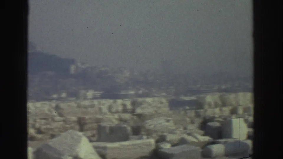 Soft 8mm view across archaeological stone remains toward a distant hillside and urban skyline.
