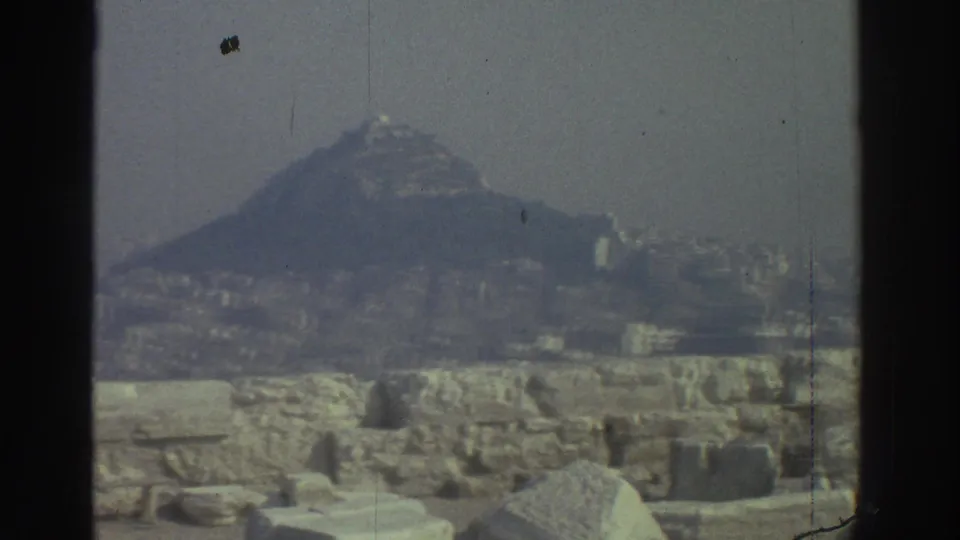 1976 home-movie frame showing a ruin foreground, distant hill, and faint urban buildings under hazy light.